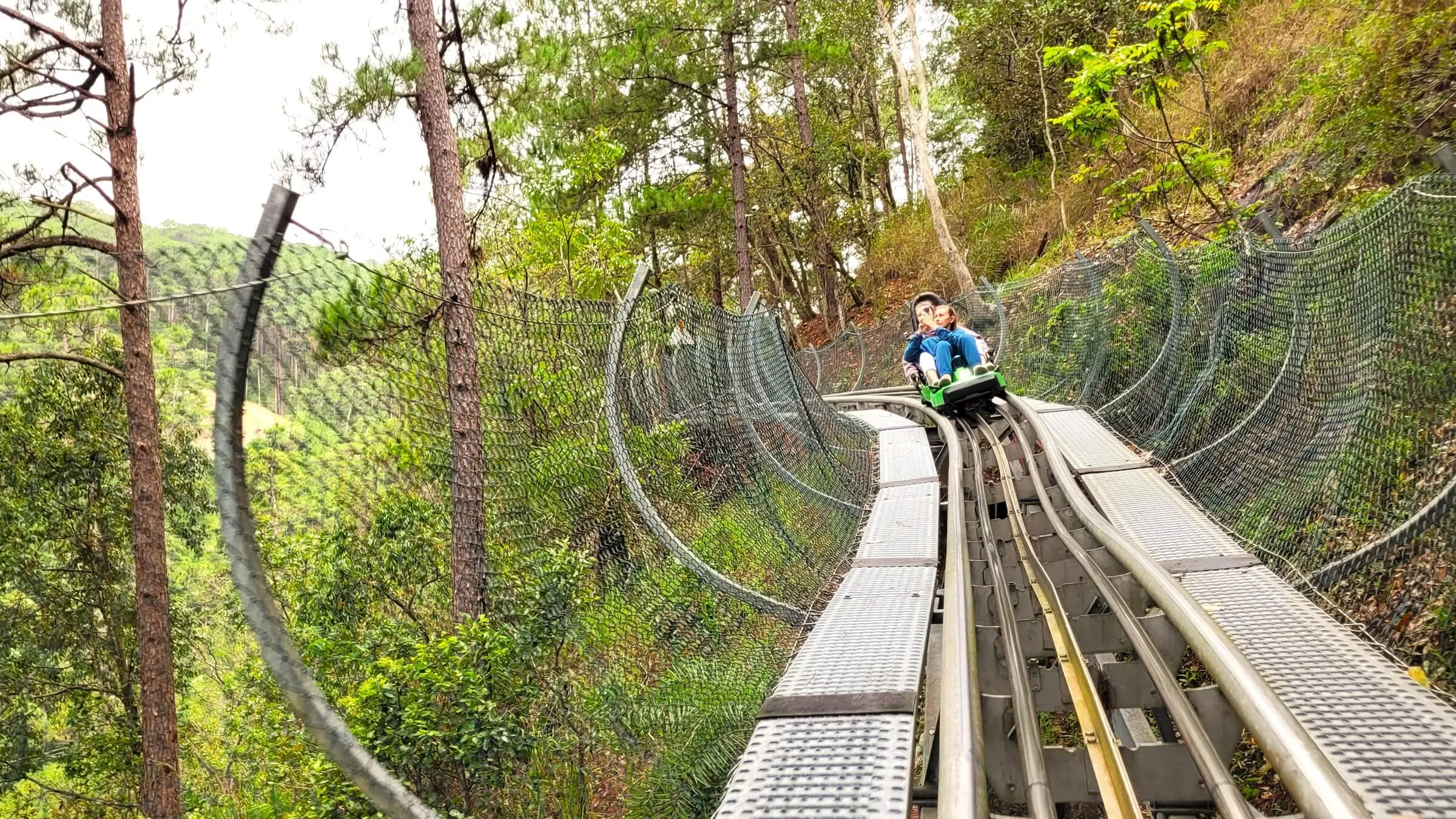 Two riders in a green car on the Da Lat Alpine Coaster, the track curving along a wooded slope above a pine valley